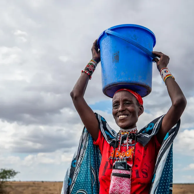 An African woman smiles while holding a large blue bucket on her head