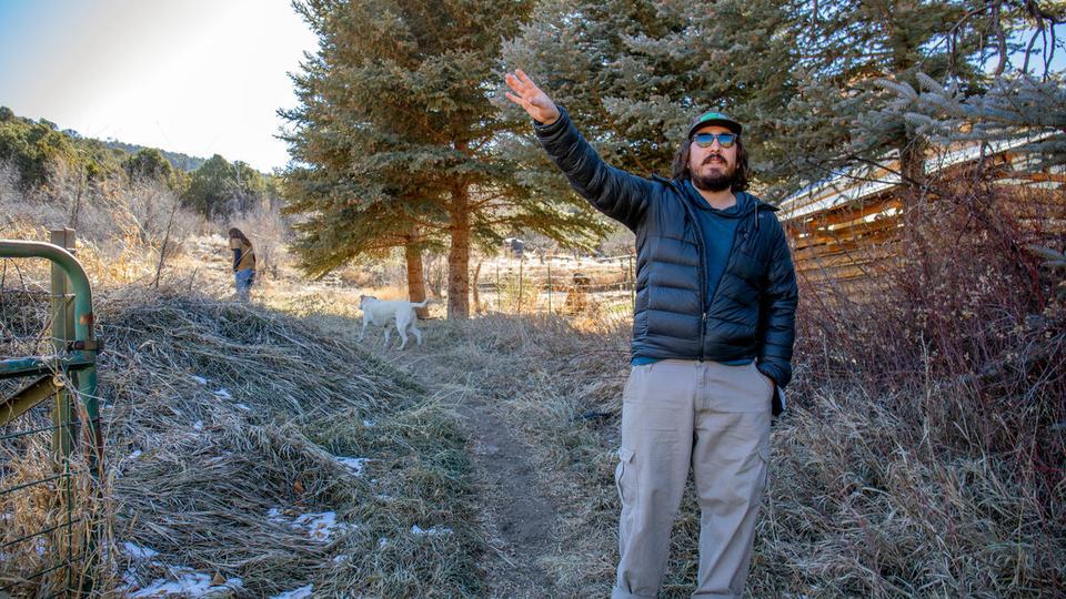 Emilio Borrego wears a navy jacket and khaki pants and gestures with his right arm standing in front of trees and hills