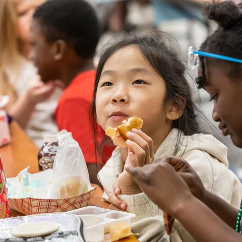Students Students Alice T. and Kyla P. eat lunch in the cafeteria at Citrus Elementary School, holding an orange and sitting at a table