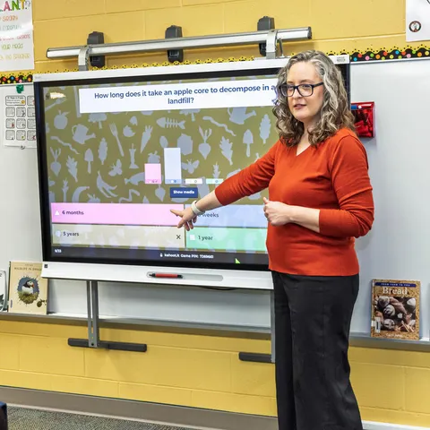 Melissa Miskovsky wears a read shirt and stands in front of a smart board in a yellow classroom using Kahoot quizzes on food waste to start a lesson on composting and food waste.