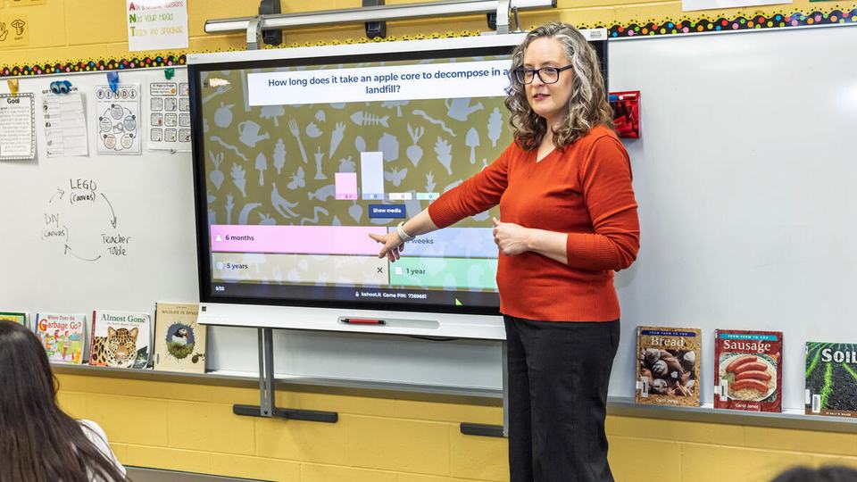 Melissa Miskovsky wears a read shirt and stands in front of a smart board in a yellow classroom using Kahoot quizzes on food waste to start a lesson on composting and food waste.