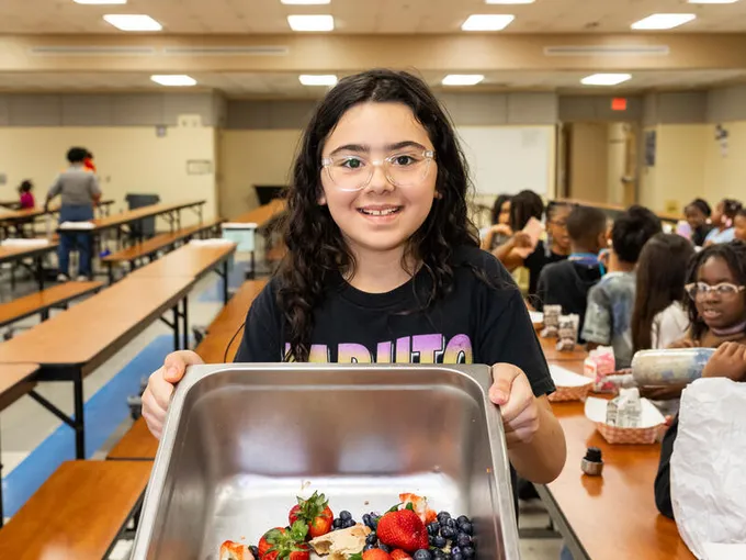 Isabelle I. has brown hair and smiles at the camera while holding up a metal bin of compostable food