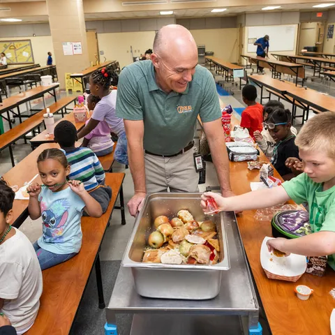 Math coach Dan St Onge wears a gray shirt and stands over a metal compost bin with students around him putting items in