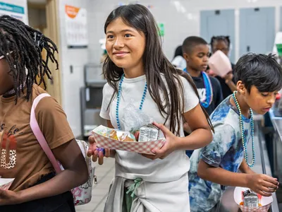 Jaden H., Sara P., Leandro R., and Carter C. stand in line in brown, gray and blue shirts, holding food they just picked up in a cafeteria