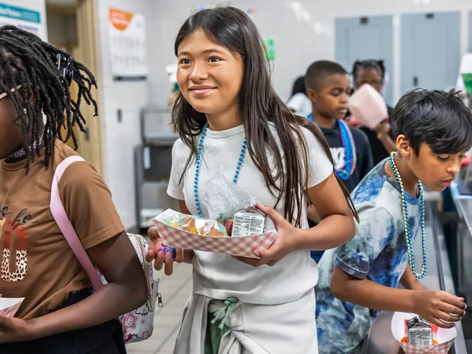 Jaden H., Sara P., Leandro R., and Carter C. stand in line in brown, gray and blue shirts, holding food they just picked up in a cafeteria