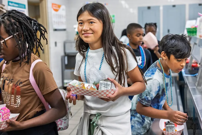 Jaden H., Sara P., Leandro R., and Carter C. stand in line in brown, gray and blue shirts, holding food they just picked up in a cafeteria