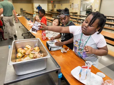 A student reaches over a cafeteria table to put waste from lunch into a collection tray.