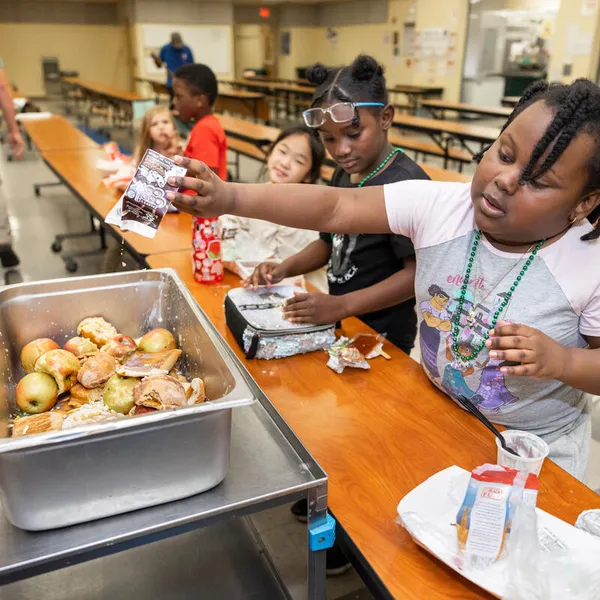 A student reaches over a cafeteria table to put waste from lunch into a collection tray.