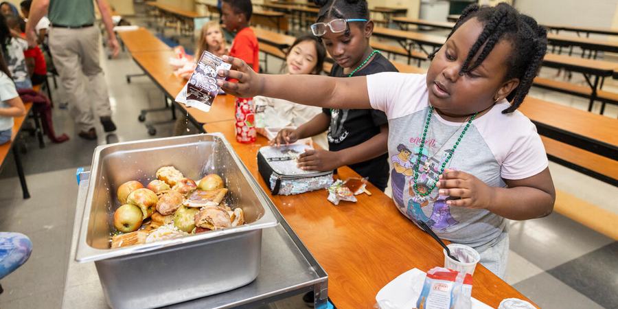 A student reaches over a cafeteria table to put waste from lunch into a collection tray.