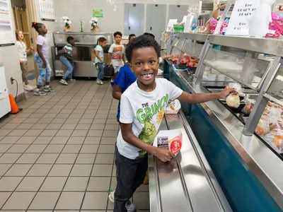 Jashiah W. wears a white shirt with a graphic and stands in a cafeteria line, grabbing lunch and smiling at the camera