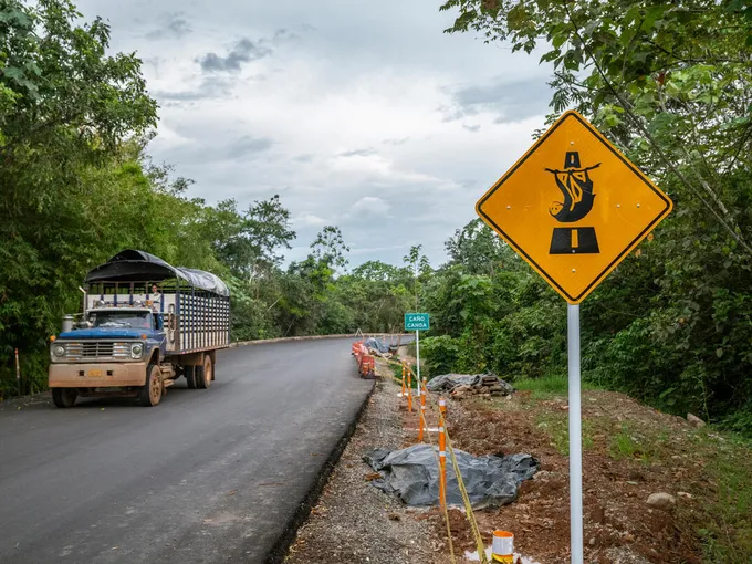 A truck travels down a road in a forest with a bright yellow sign with an image of a sloth crossing a road