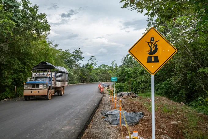 A truck travels down a road in a forest with a bright yellow sign with an image of a sloth crossing a road