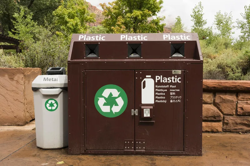 A large brown recycling dumpster next to a small gray recycling bin in front of a stone wall and greenery