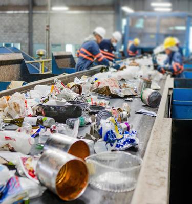 A conveyor belt moves recycling toward a team of people in blue jackets with orange stripes in a warehosue