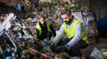 Two people wearing bright yellow vests stand among piles of recycling, separating the pieces
