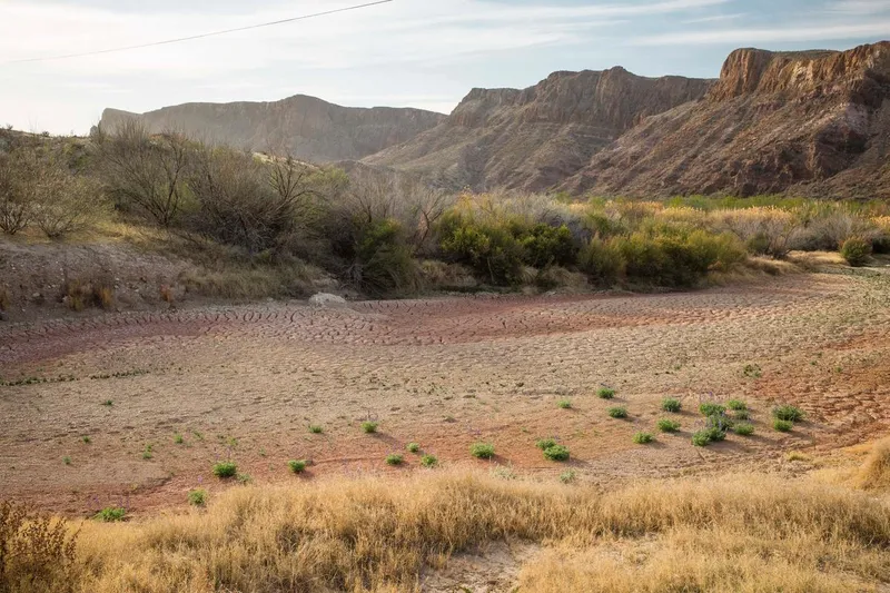 A dry river bed near the Rio Grande with brown dirt, tan grasses, and tall, rocky red cliffs in the background