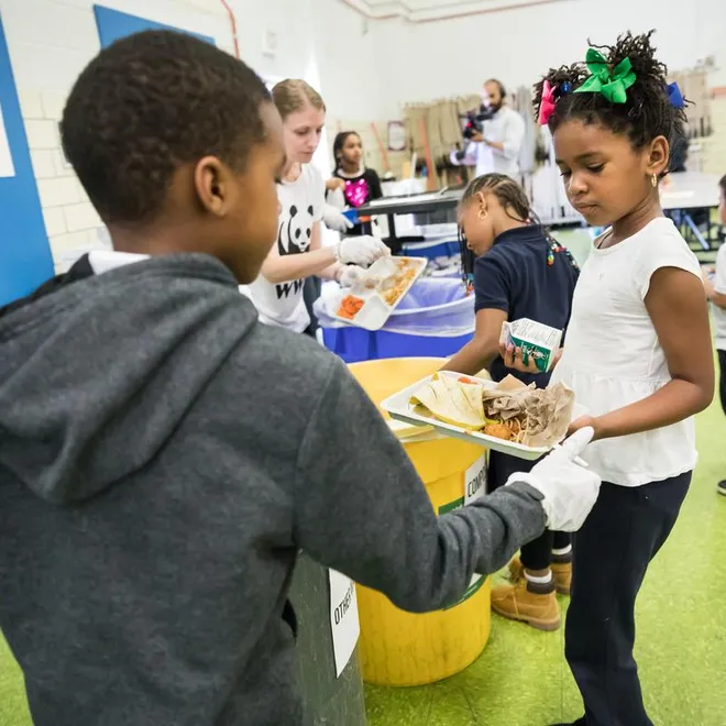 A student in a gray sweatshirt collects uneaten food from a student in a white shirt with a green bow in a cafeteria