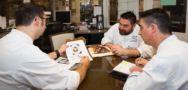 Three chefs sit around table looking at materials