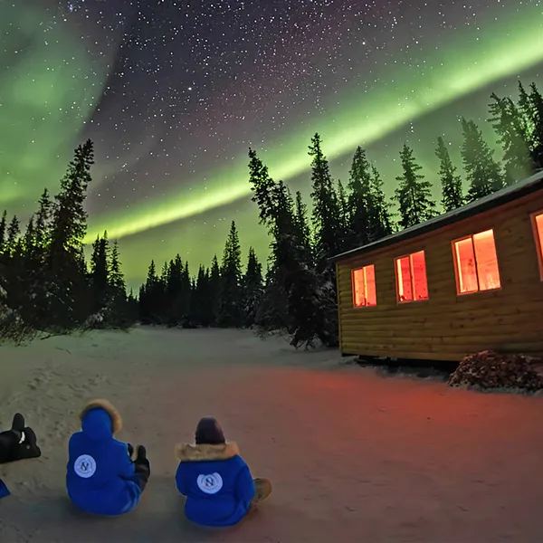 Three people in blue jackets sitting ont he ground outside a cabin, looking up at the aurora borealis lights in the night sky