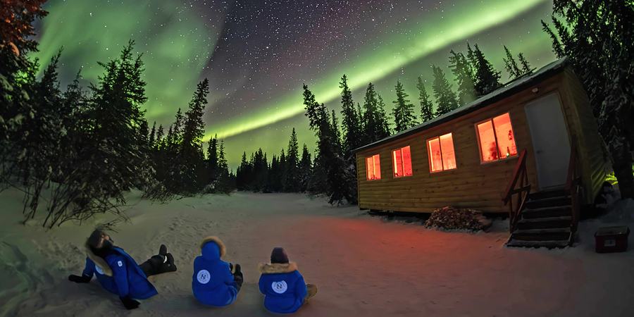 Three people in blue jackets sitting ont he ground outside a cabin, looking up at the aurora borealis lights in the night sky