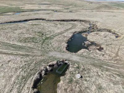 An aerial view of tan grasslands with a dark river winding through it and a metal fence-like structure built in the waterway