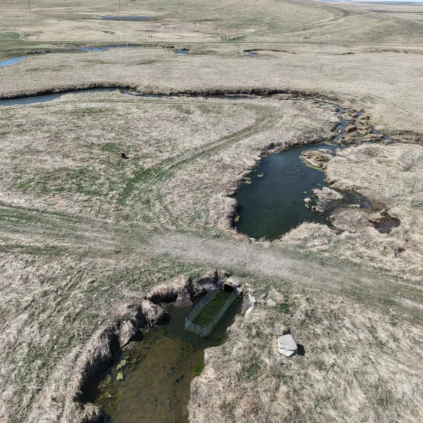 An aerial view of tan grasslands with a dark river winding through it and a metal fence-like structure built in the waterway
