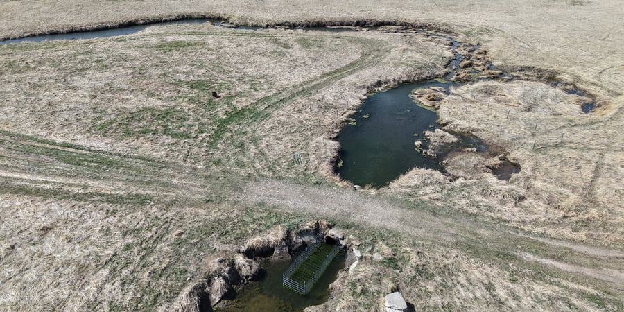 An aerial view of tan grasslands with a dark river winding through it and a metal fence-like structure built in the waterway