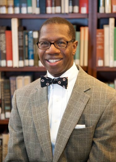 A Black man with dark-framed glasses smiles. He is wearing a checkered gray suit, white shirt, and black bow-tie with patterns.