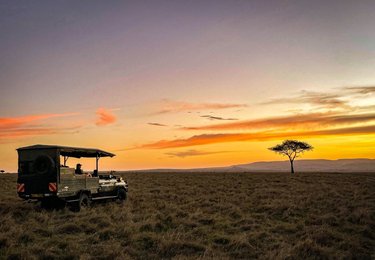 Safari truck sits on the left of the frame in front of wide landscape and a single tree silhouetted against an orange sky at sunset