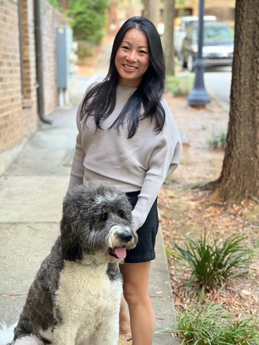 Stephanie Zhu stands on the sidewalk in black shorts and a gray shirt with a gray and white dog.