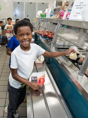 Jashiah W. wears a white shirt and a big grin on his face as he reaches for food in a cafeteria line.