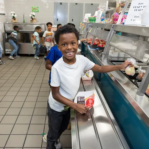 Jashiah W. wears a white shirt and a big grin on his face as he reaches for food in a cafeteria line.