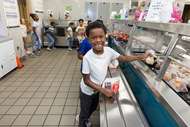 Jashiah W. wears a white shirt and a big grin on his face as he reaches for food in a cafeteria line.