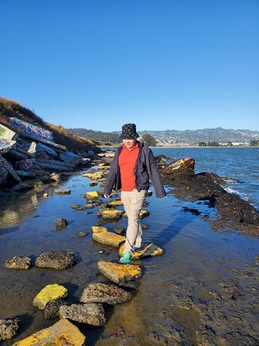 Peter Pham wears a red shirt and a hat and walks along a shore on a sunny day