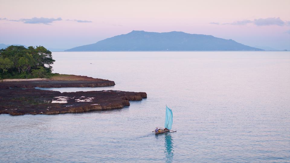 A small sailboat with a blue sail floats near shore, with another island in the distance.