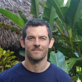 A headshot of Henry Harrison, who has brown hair and a navy blue shirt and stands in front of greenery