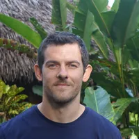 A headshot of Henry Harrison, who has brown hair and a navy blue shirt and stands in front of greenery