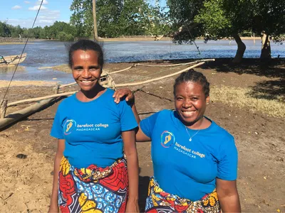 Two smiling women stand next to each other on the muddy bank of a wide river