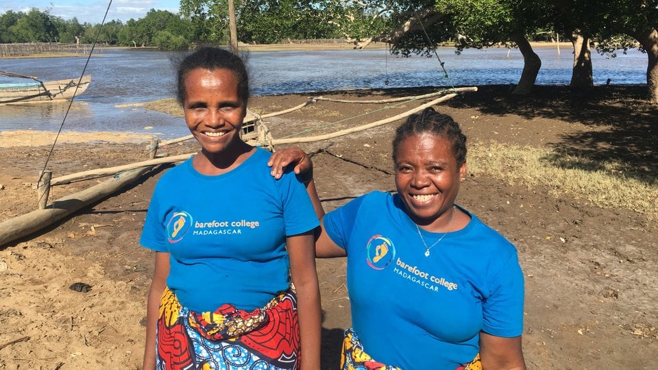 Two smiling women stand next to each other on the muddy bank of a wide river
