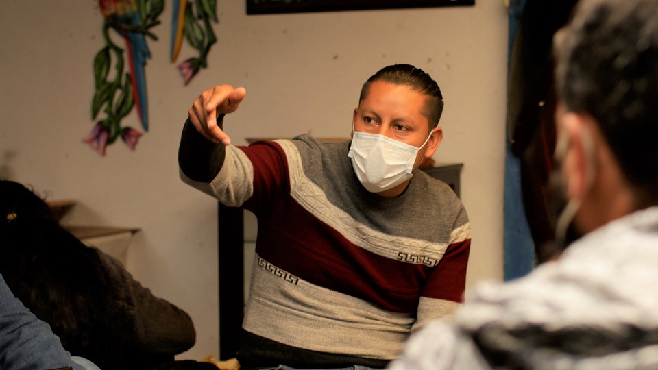 Man with white medical mask on speaks to a group of people all sitting down
