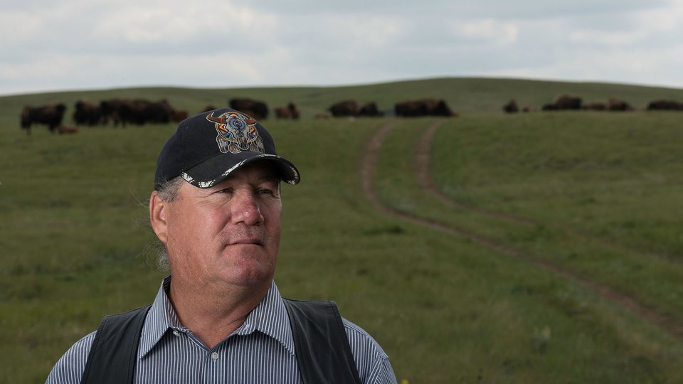Robbie Magnan wears a black baseball hat standing in front of a dirt road in a field