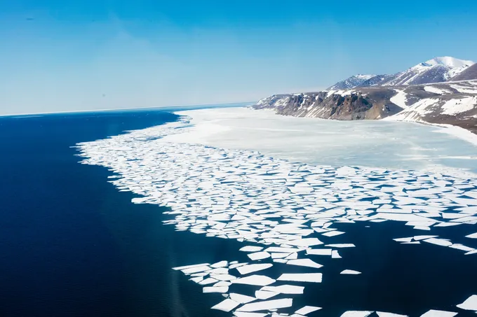 An aerial view of chunks of broken ice alongside a barren icy coast and a blue ocean