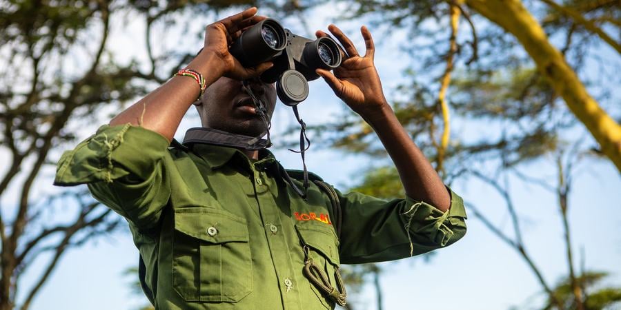 A SORALO community ranger in Kenya wears an olive green uniform and looks through a pair of binoculars with trees and sky in the background