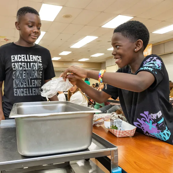Ruben P. wears a black shirt and pushed a cart with a metal bin on top collecting food waste from another student, Myles B., at a table