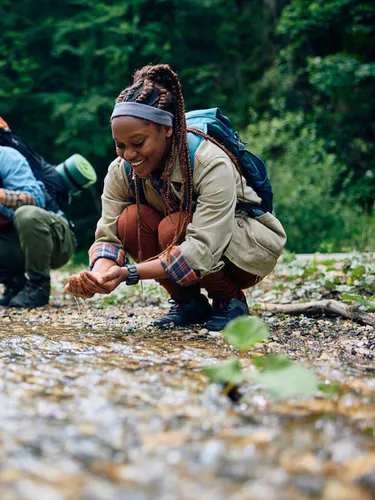 Low angle shot of 2 people with hiking gear bending down at stream scooping up water with hands. Smiling, connecting with nature. Trees in the background.