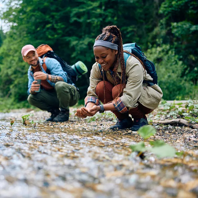 Low angle shot of 2 people with hiking gear bending down at stream scooping up water with hands. Smiling, connecting with nature. Trees in the background.