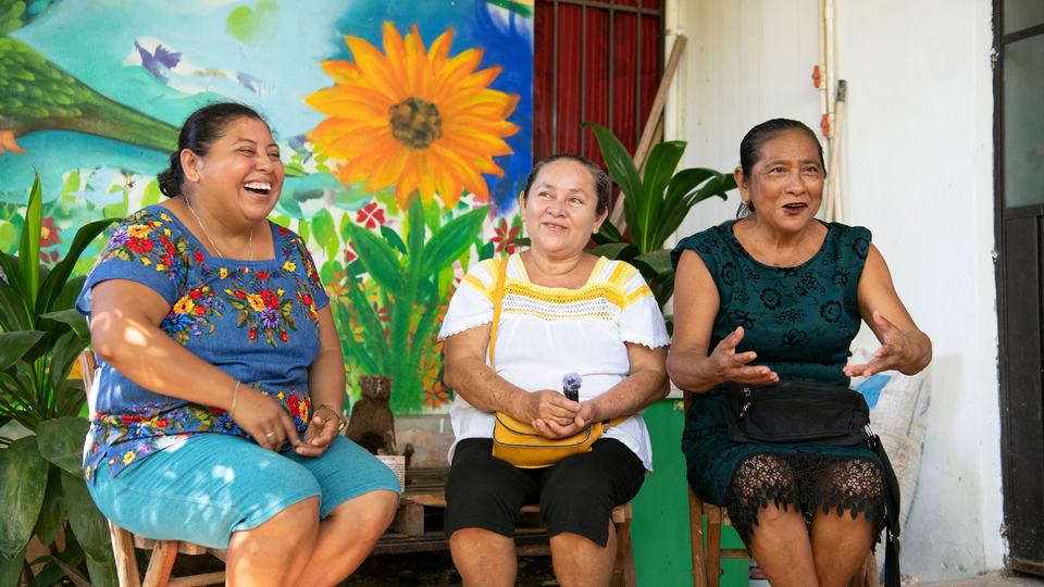 Three ladies smiling, sitting in front of colorful mural