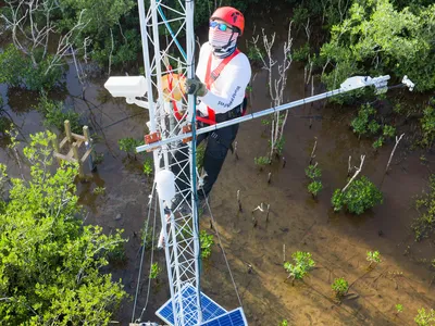 Aerial photo of a man in a helmet and mask on a white radio tower, checking equipment