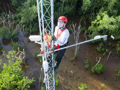 Aerial photo of a man in a helmet and mask on a white radio tower, checking equipment