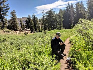 Dr. Cody Aylward wears a hat and dark clothing and kneels on a small dirt path in an open area to collect a scat sample with trees in the background